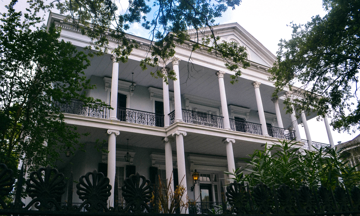 A picture of a mansion nestled between trees. It has white columns and black railings. 